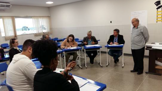 Fotografia de uma sala de aula com pessoas sentadas em carteiras escolares, que estão organi-zadas em um círculo. Ao lado há um homem em pé.